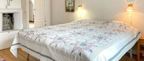 A bedroom with a large bed, a fireplace, two wall-mounted lamps, and a door leading to another room.