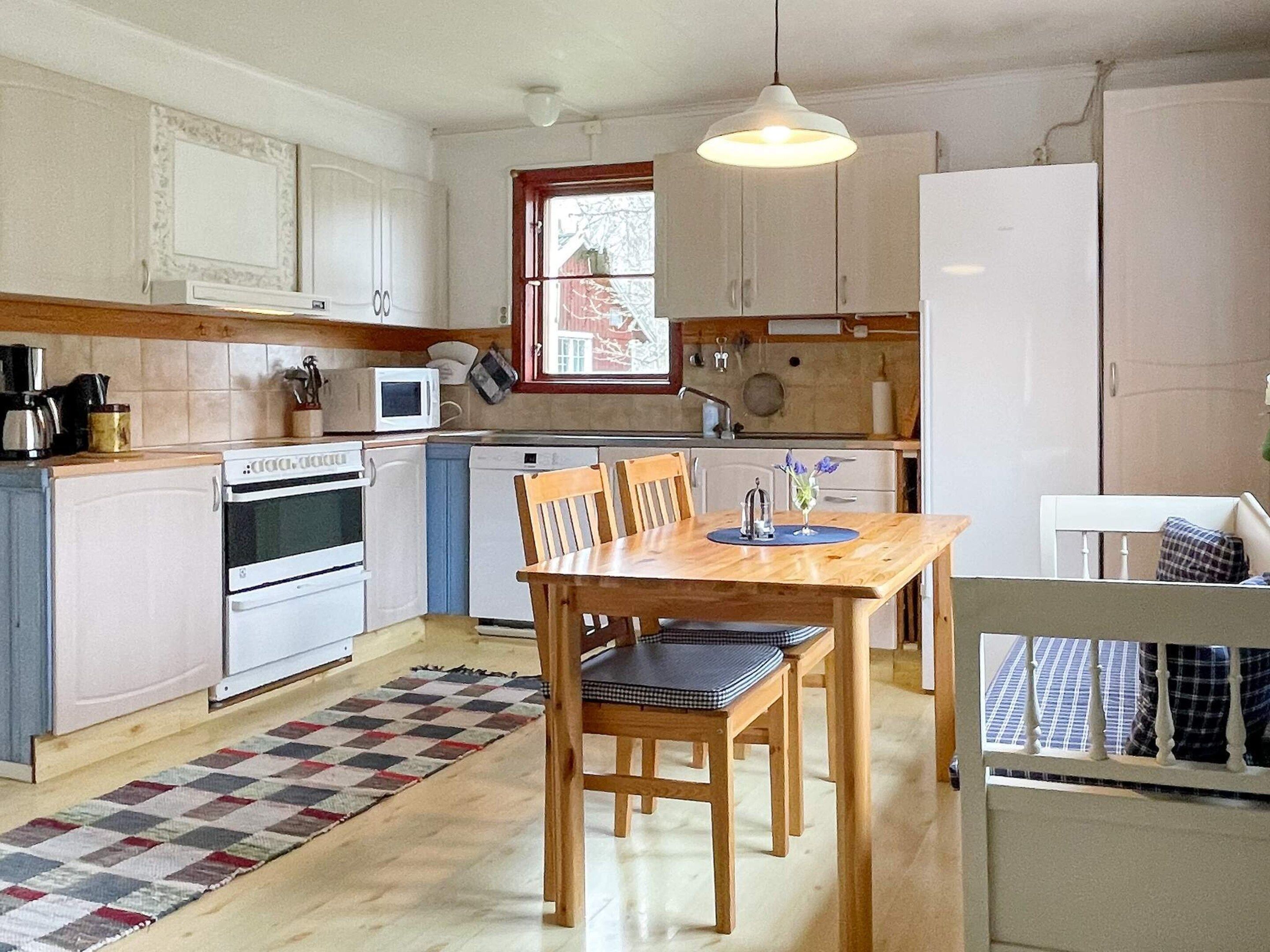 Een kleine keuken met witte apparaten, een houten eettafel en een geruit tapijt.