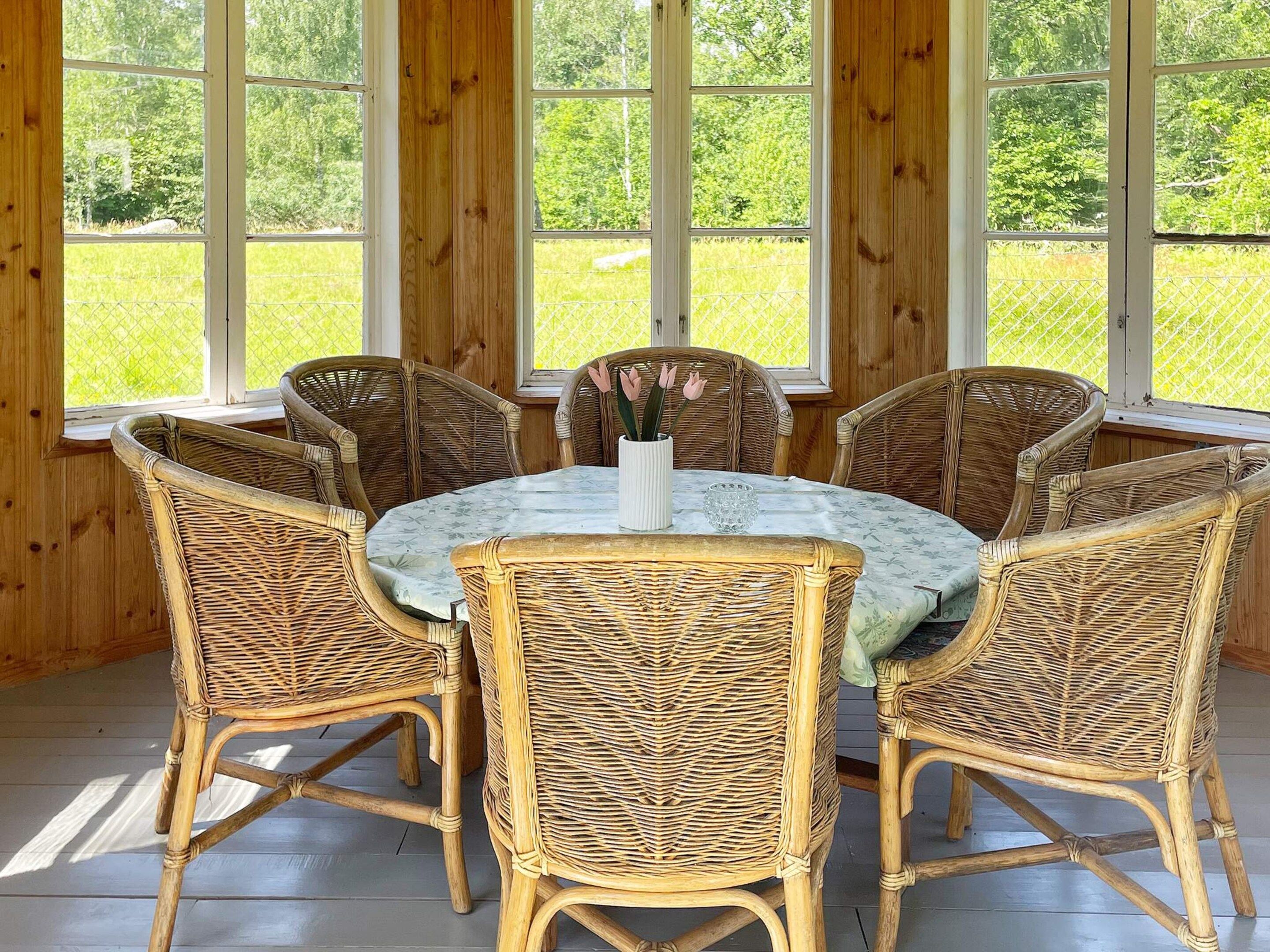 A dining area with a round table and six wicker chairs, large windows, and a view of a green landscape.