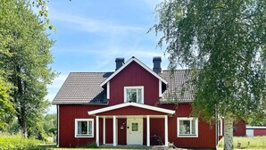 Ein rotes, zweistöckiges Haus mit weißem Balkon, umgeben von viel Grün und blauem Himmel.