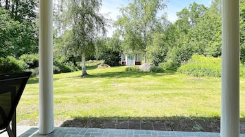 Een overdekt terras met uitzicht op een gazon en een klein gebouw in de verte.