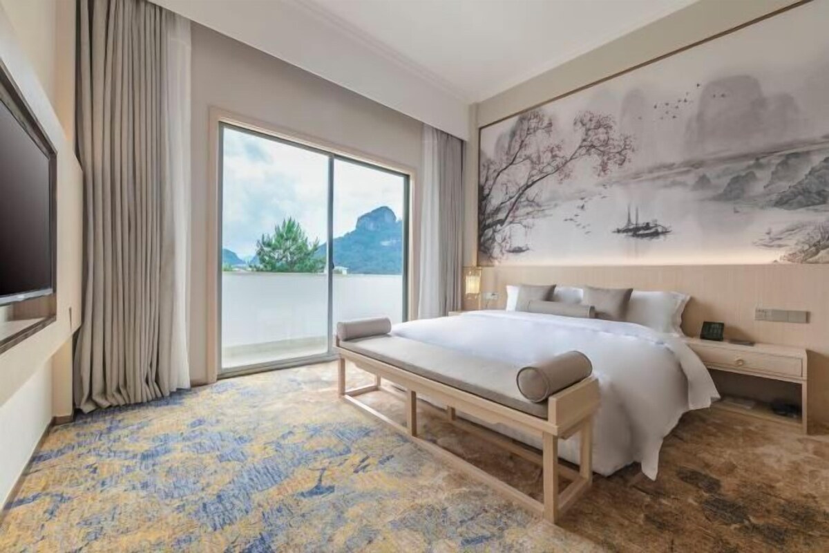 A hotel room with a large bed, a bench, a desk, a flat-screen TV, and a painting on the wall.