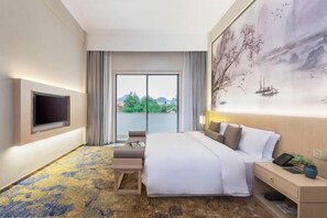 A hotel room with a bed, a desk, a TV, a large window, and a painting on the wall.