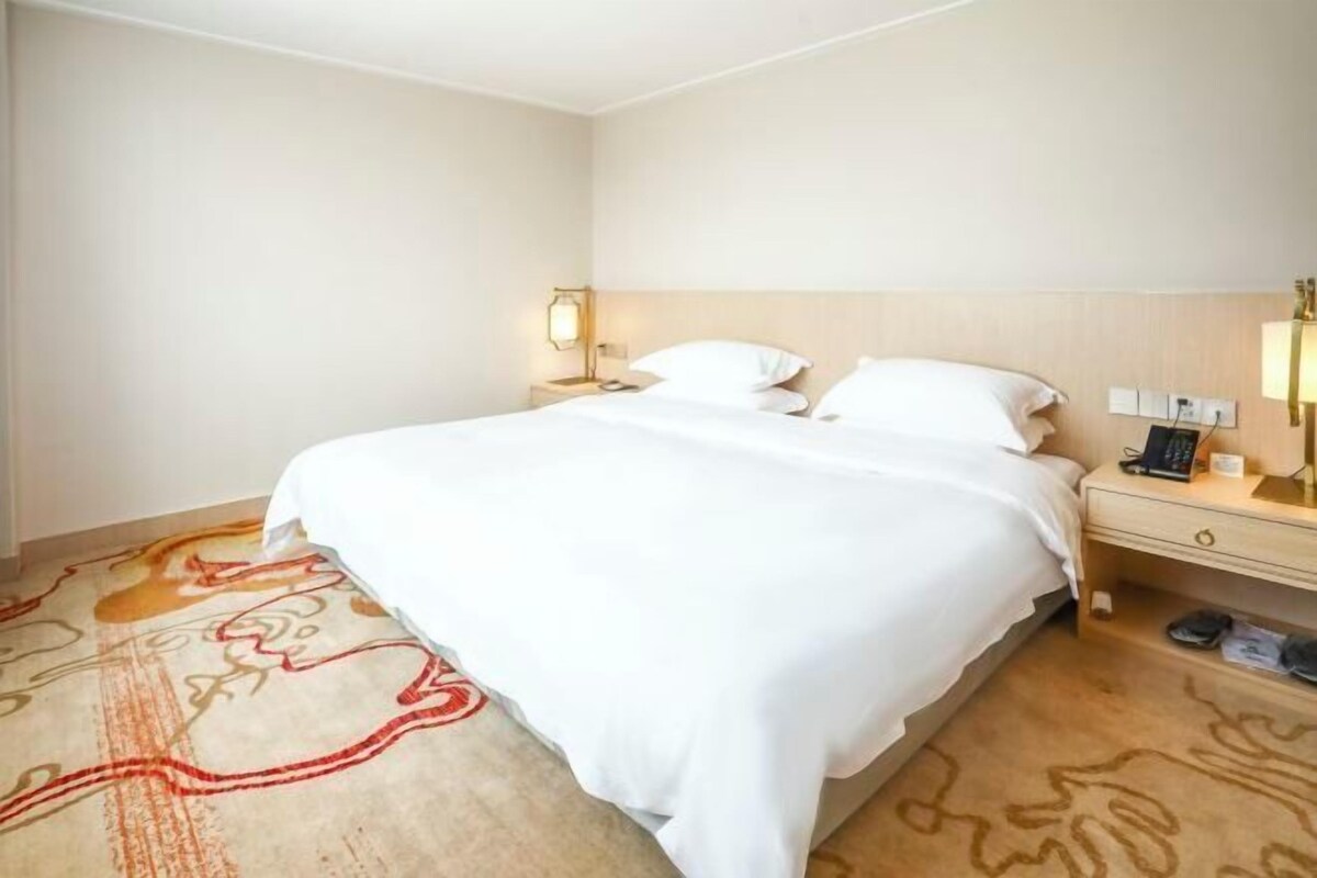 A hotel room with a large bed, a bedside table with a lamp, and a carpet with a red design.