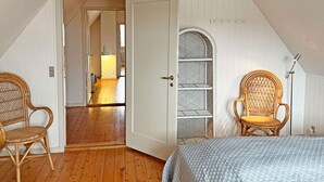 A bedroom with a sloped ceiling, wooden flooring, a bed with a blue quilt, two wicker chairs, a white door, and a small shelf unit. - 6 Person Holiday Home in Svaneke (Svaneke)