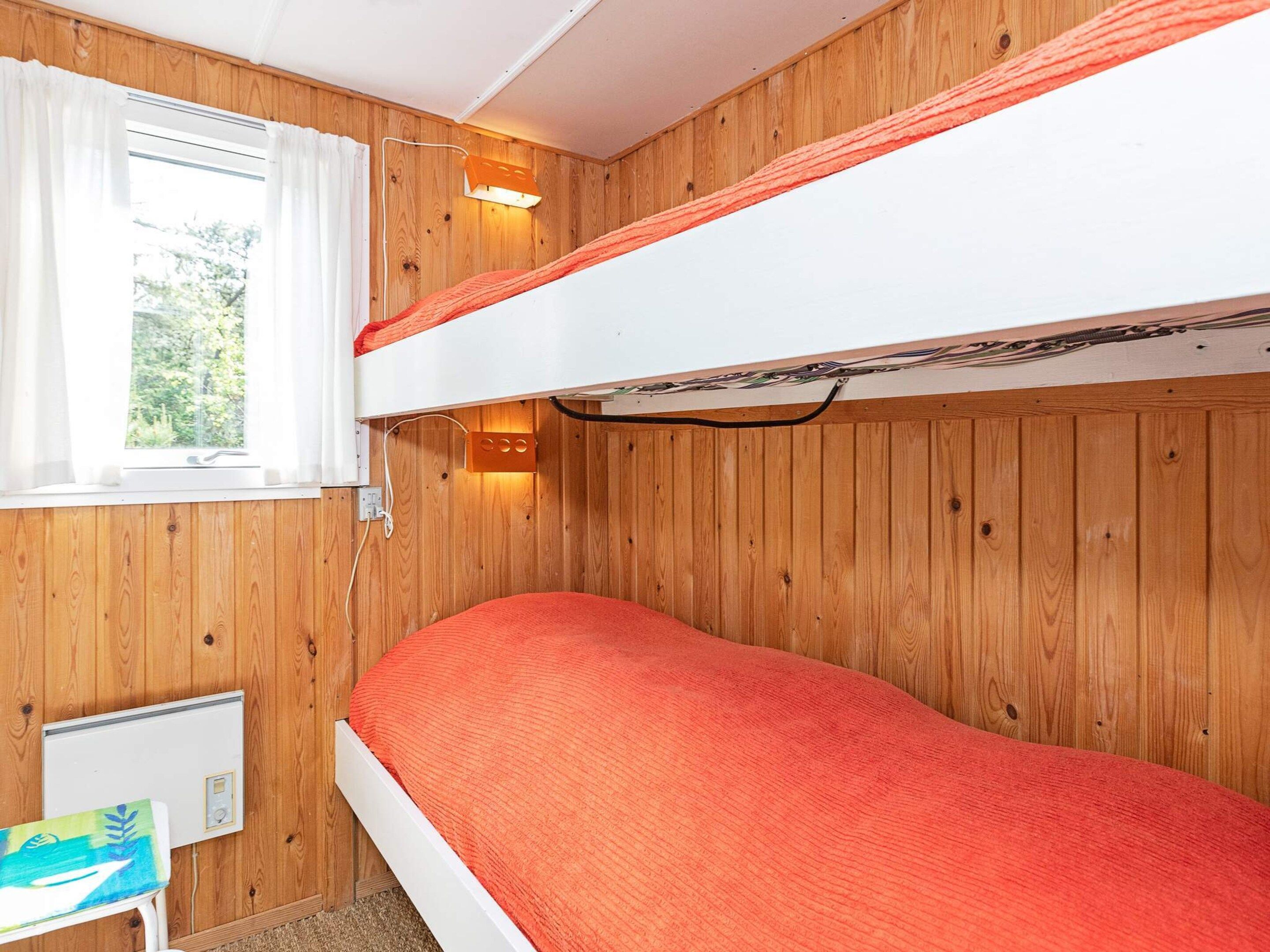 A bunk bed with a red mattress in a room with wooden walls and a window with white curtains.