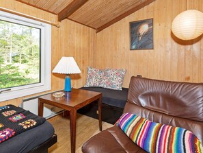 A cozy room with a wooden ceiling, a window with a view of greenery, a brown leather sofa, a small wooden table with a lamp, and a colorful cushion. - 4 Person Holiday Home in Blokhus (Blokhus)