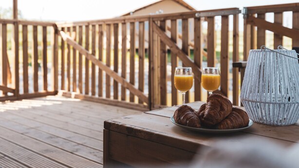 Una terraza de madera con una mesa dispuesta para el desayuno, que incluye cruasanes y vasos de zumo de naranja.