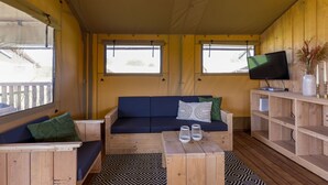 Interior acogedor de una cabaña con suelo de madera, un sofá azul con cojines, un televisor sobre una estantería de madera y una ventana con vistas al exterior.