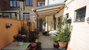 Eine schmale Außenpatio mit Tisch und Stühlen, umgeben von Topfpflanzen und einer Pergola.