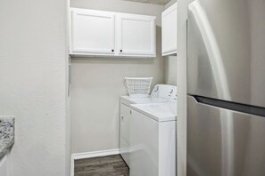 A compact laundry room with a front-loading washer and dryer, a stainless steel refrigerator, and white cabinetry above the appliances. - I043 - Cozy 2BD | Pool | DFW South 183/161 (Irving)