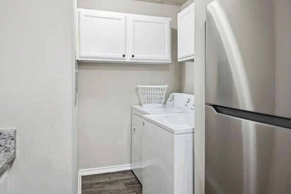 A compact laundry room with a front-loading washer and dryer, a stainless steel refrigerator, and white cabinetry above the appliances.