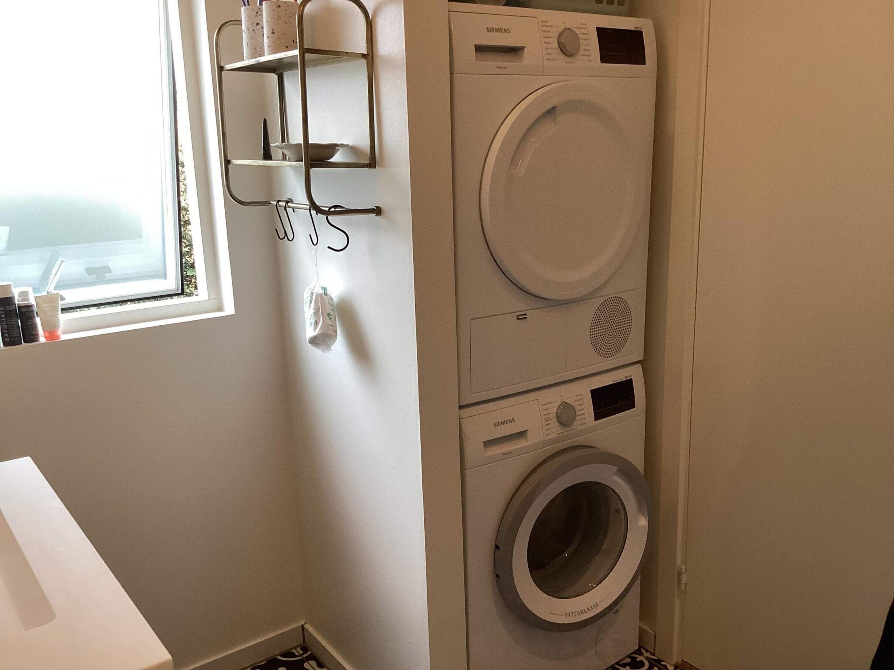 A laundry area with a stacked washer and dryer, a window, and a metal shelf with cups.