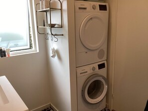 A laundry room with a stacked washer and dryer, a window, and a metal shelf with cups. - 4 Star Holiday Home in Juelsminde (Juelsminde)