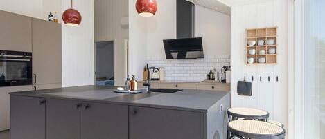Una cocina moderna con una isla central, horno empotrado y un televisor grande sobre la estufa. Hay dos lámparas colgantes y un estante de madera con utensilios de cocina.