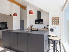 A modern kitchen with a central island, a built-in oven, and a large TV above the stove. There are two hanging pendant lights and a wooden shelf with kitchenware. - 4 Star Holiday Home in Juelsminde (Juelsminde)