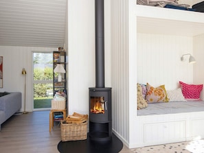 Una sala de estar acogedora con un estufa de leña negra, una estantería, una cesta de mimbre y un asiento junto a la ventana con cojines de colores.