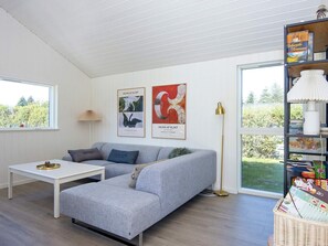 A modern living room with a grey sectional sofa, a white coffee table, and a floor lamp. There are two framed artworks on the wall, a window with a view of greenery, and a bookshelf with various items.