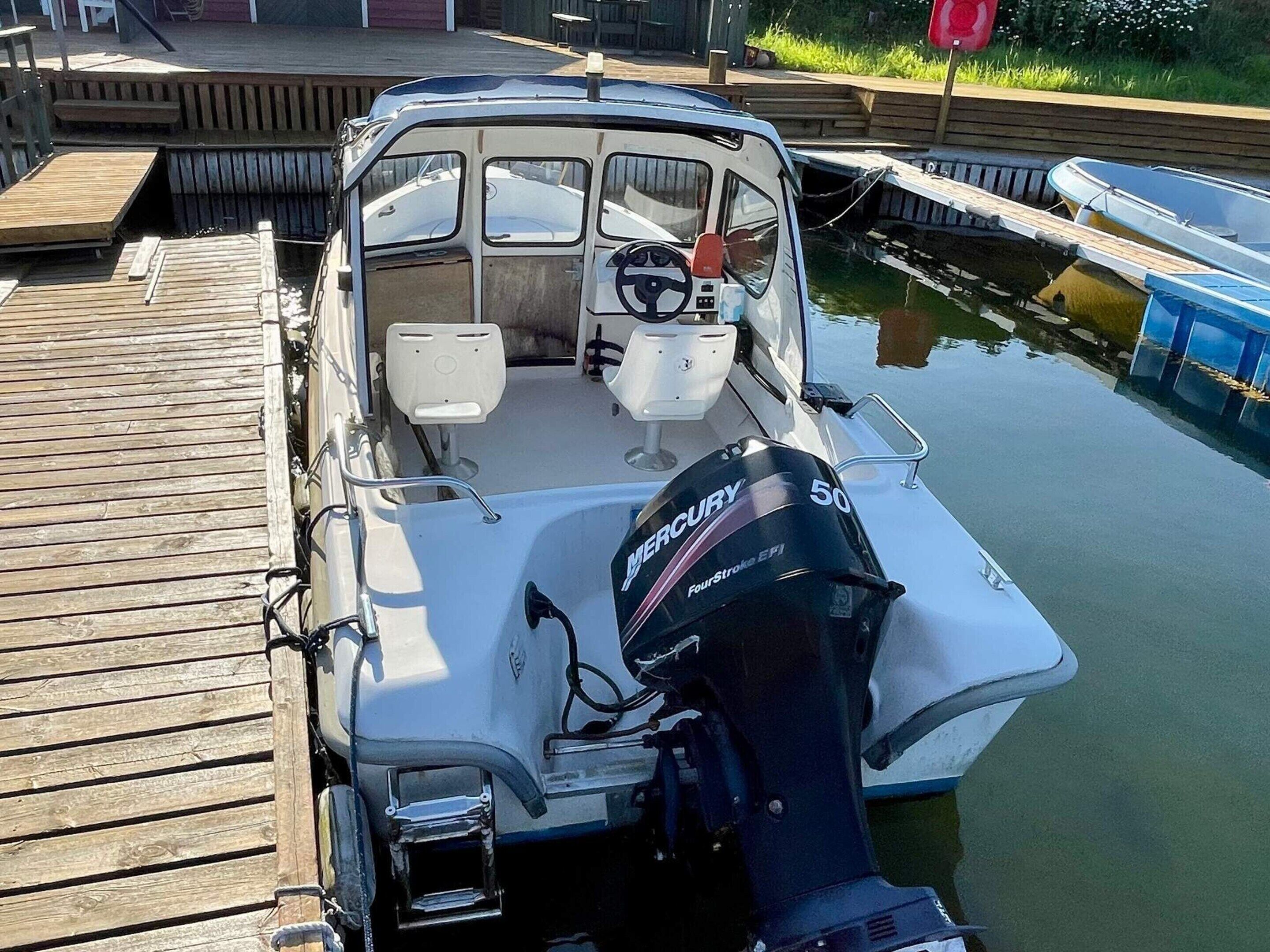 En lille motorbåd ligger til kaj ved en træbrygge, monteret med en Mercury 50 påhængsmotor.