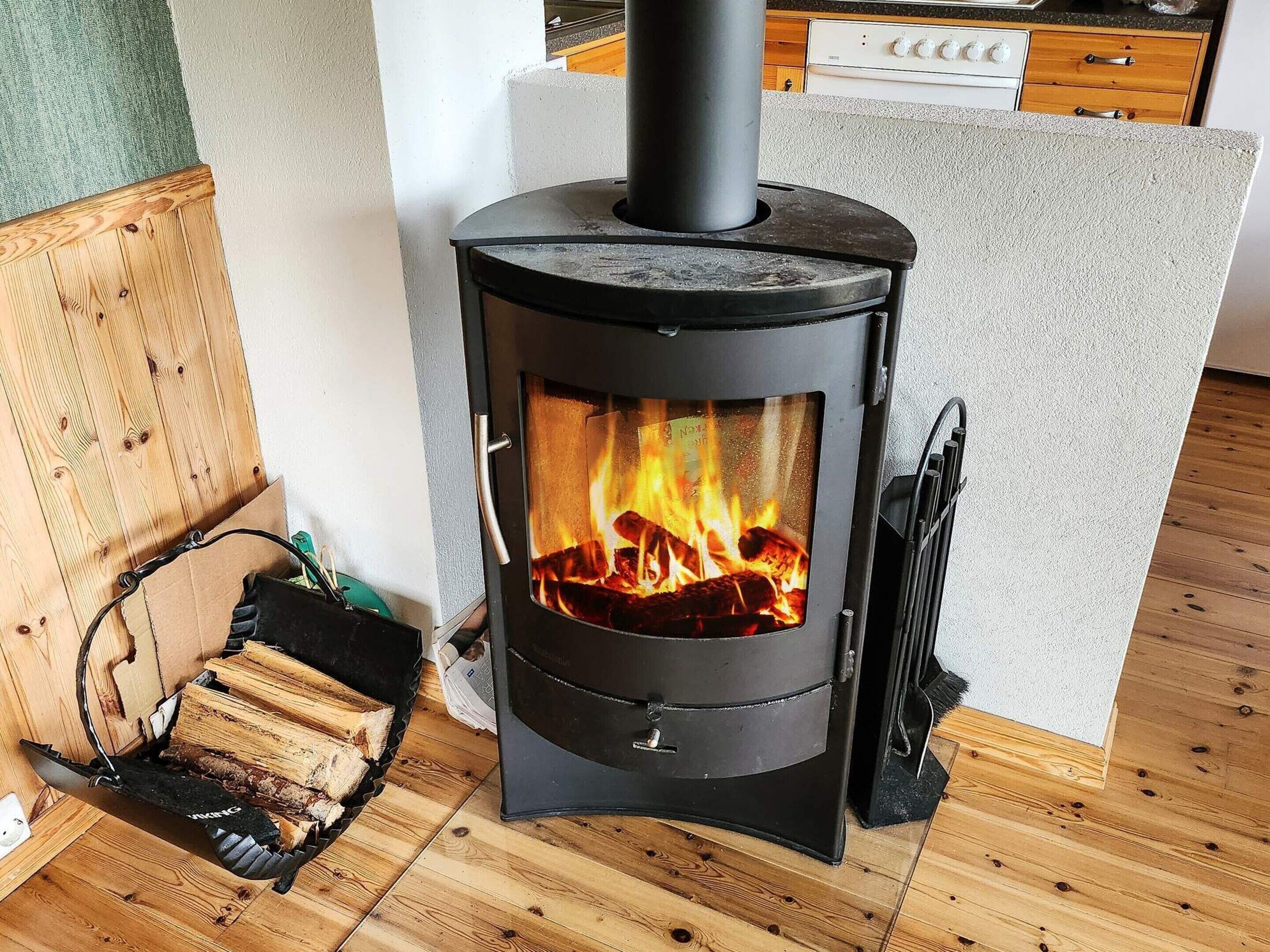 Un poêle à bois avec des flammes apparentes, un espace de rangement pour le bois et une cuisine en arrière-plan.