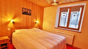 Un dormitorio con cabecera de madera, cama con ropa de cama blanca, mesita de noche con cajón, lámpara de pared y ventana con adornos con forma de corazón.