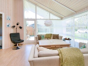 Una sala de estar moderna con sofá, una mesita de centro y una silla. Hay un ventanal con persianas, una puerta que da al exterior y un ventilador de techo.