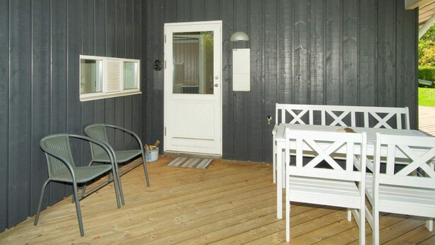 Un espace extérieur abrité doté d’une terrasse en bois, d’une porte blanche, de deux chaises en métal et d’un banc blanc.