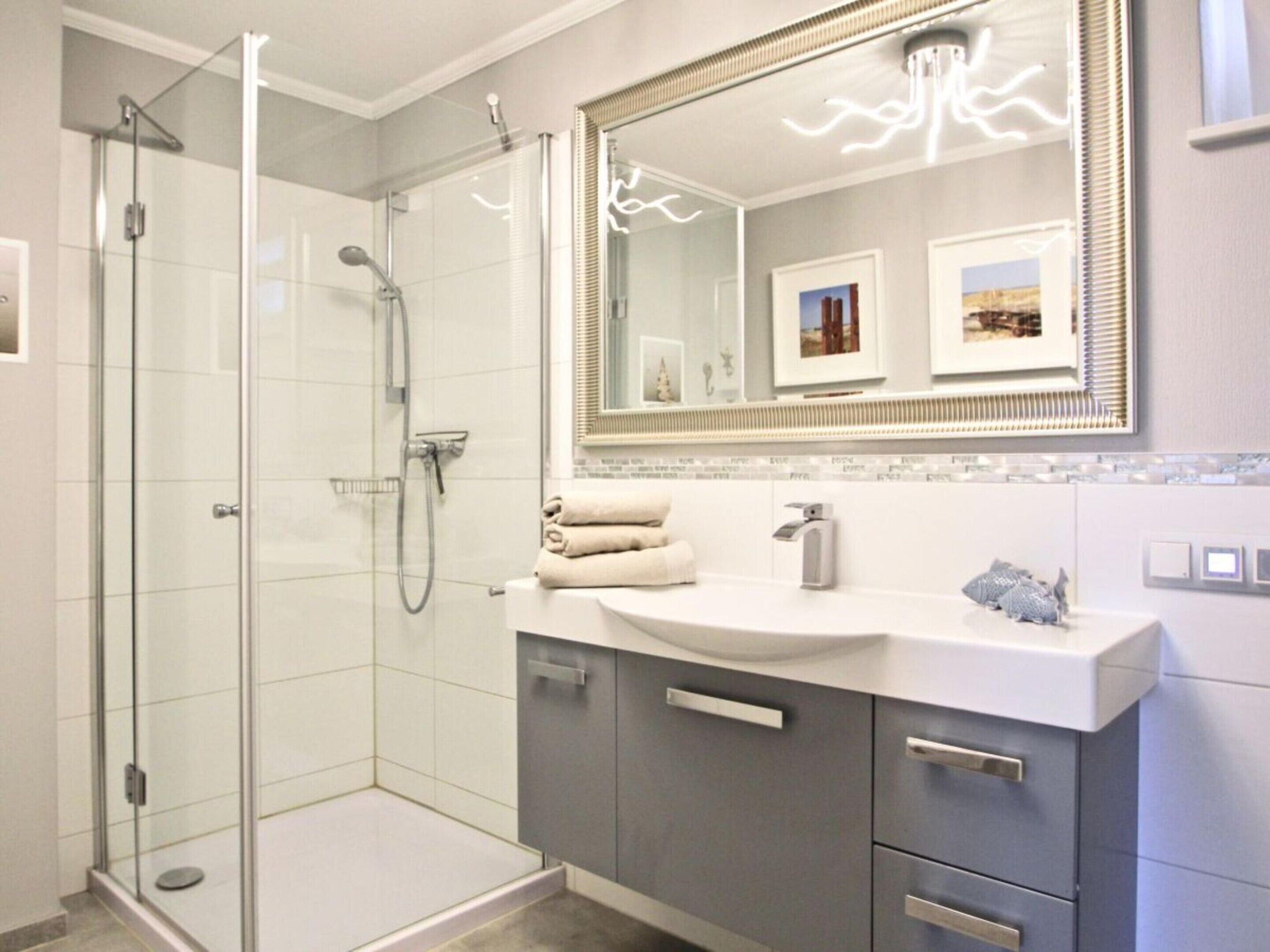 A modern bathroom with a glass shower enclosure, a large mirror with decorative lights, and a white vanity with grey cabinets.