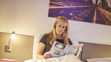 A person sitting on a bed reading a magazine. There is a nightstand with a lamp and a book on it. A cityscape painting is on the wall behind the person.