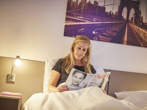 A person sitting on a bed reading a magazine. There is a nightstand with a lamp and a book on it. A cityscape painting is on the wall behind the person. - Great Villa With Luxurious Terrace, Near Roermond (Roggel)