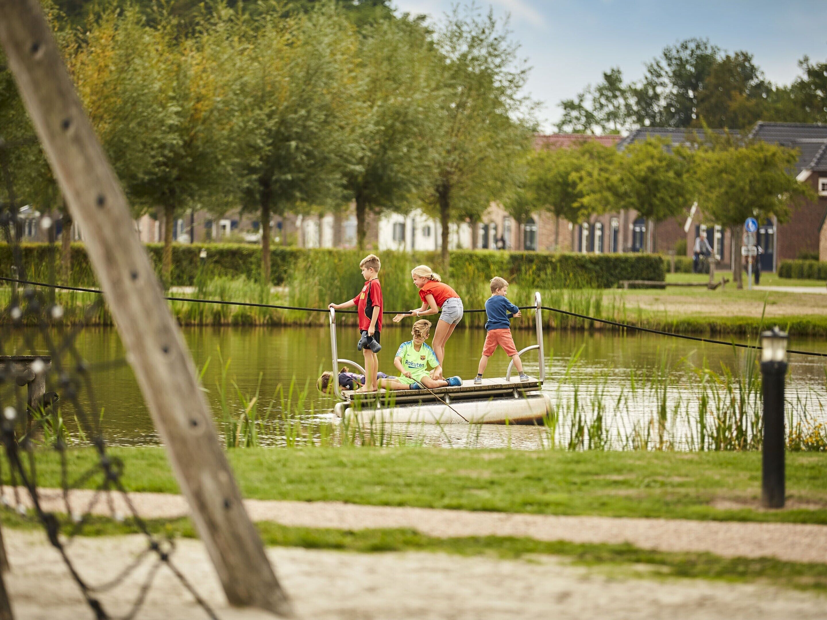 Eine Gruppe Kinder spielt auf einem kleinen Boot in ruhigem Gewässer, umgeben von viel Grün und einigen Häusern im Hintergrund.