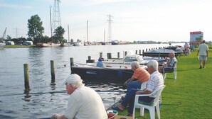 Uma marina com barcos atracados, uma área gramada com pessoas sentadas em cadeiras e uma placa de sinalização ao fundo.