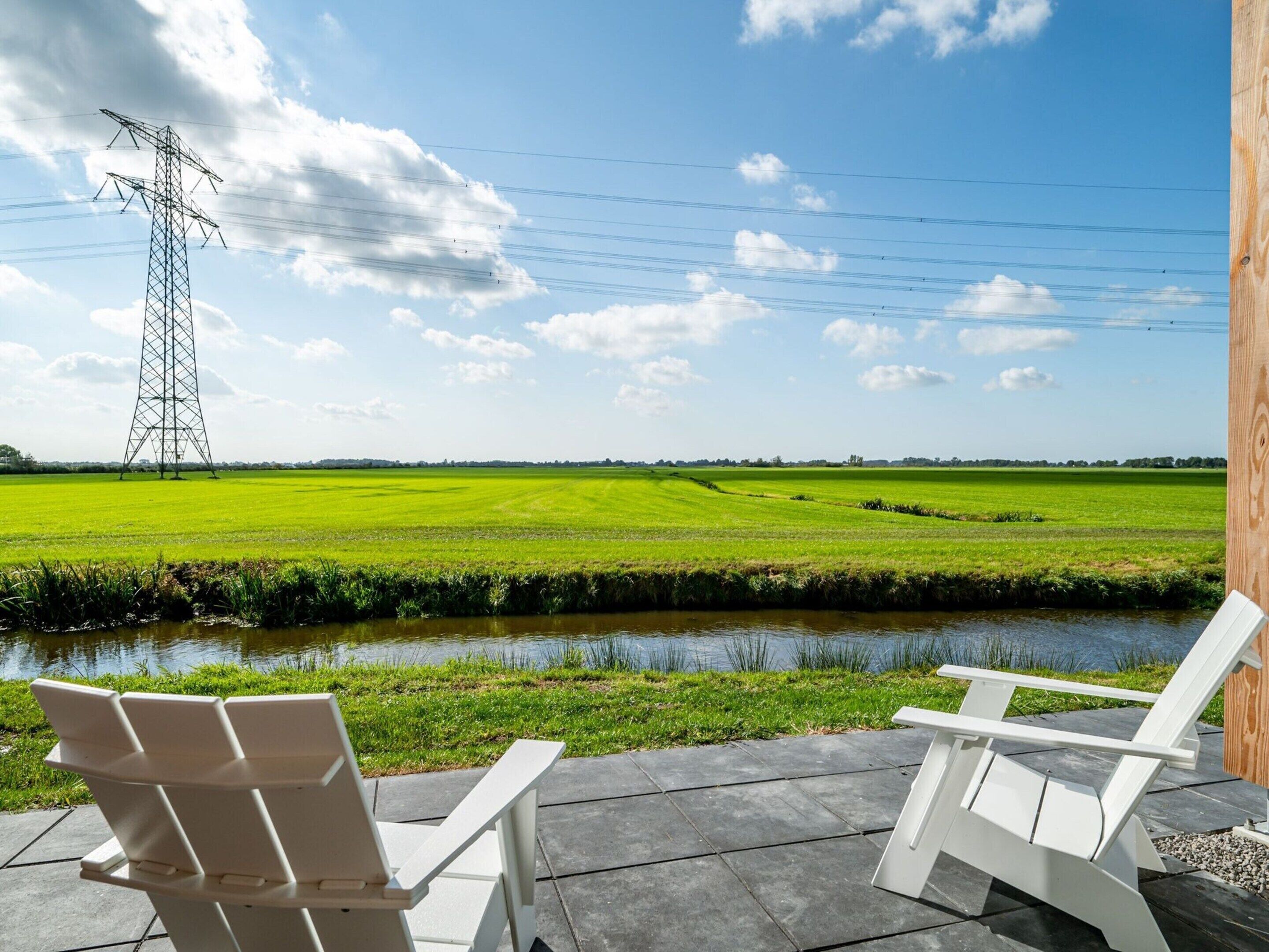 A view of a green field with a power line tower in the distance, a canal, and two white chairs on a patio.
