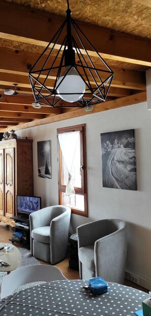A room with a wooden ceiling, a modern pendant light, two grey armchairs, a round table with a polka dot tablecloth, and a television on a wooden cabinet.