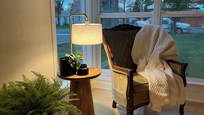 Ein gemütliches Zimmer mit Holzboden, einem Sessel mit weißer Decke, einer Lampe und einer Topfpflanze.