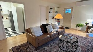 Ein Wohnzimmer mit einem braunen Ledersofa, Teppichen mit Muster, einem runden Couchtisch und einer weißen Tür, die in eine Küche führt.