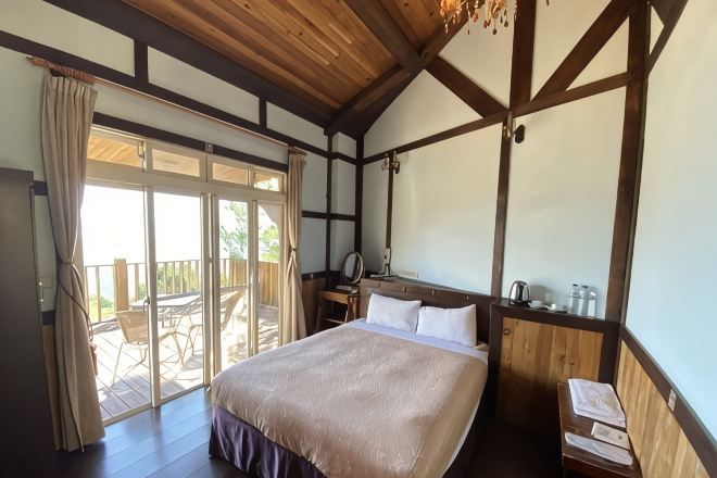 A bedroom with a large bed, wooden ceiling beams, a nightstand, a mirror, and a view of the outdoors.