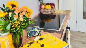 Una cocina con armarios naranjas, una mesa de vidrio, un florero con flores, una cesta de frutas, un libro y una botella.