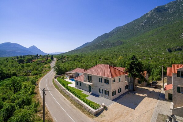 A two-story house with a red-tiled roof, surrounded by lush greenery and a winding road. - Villa in Croatia With Stunning Mountain Views (Vrgorac)