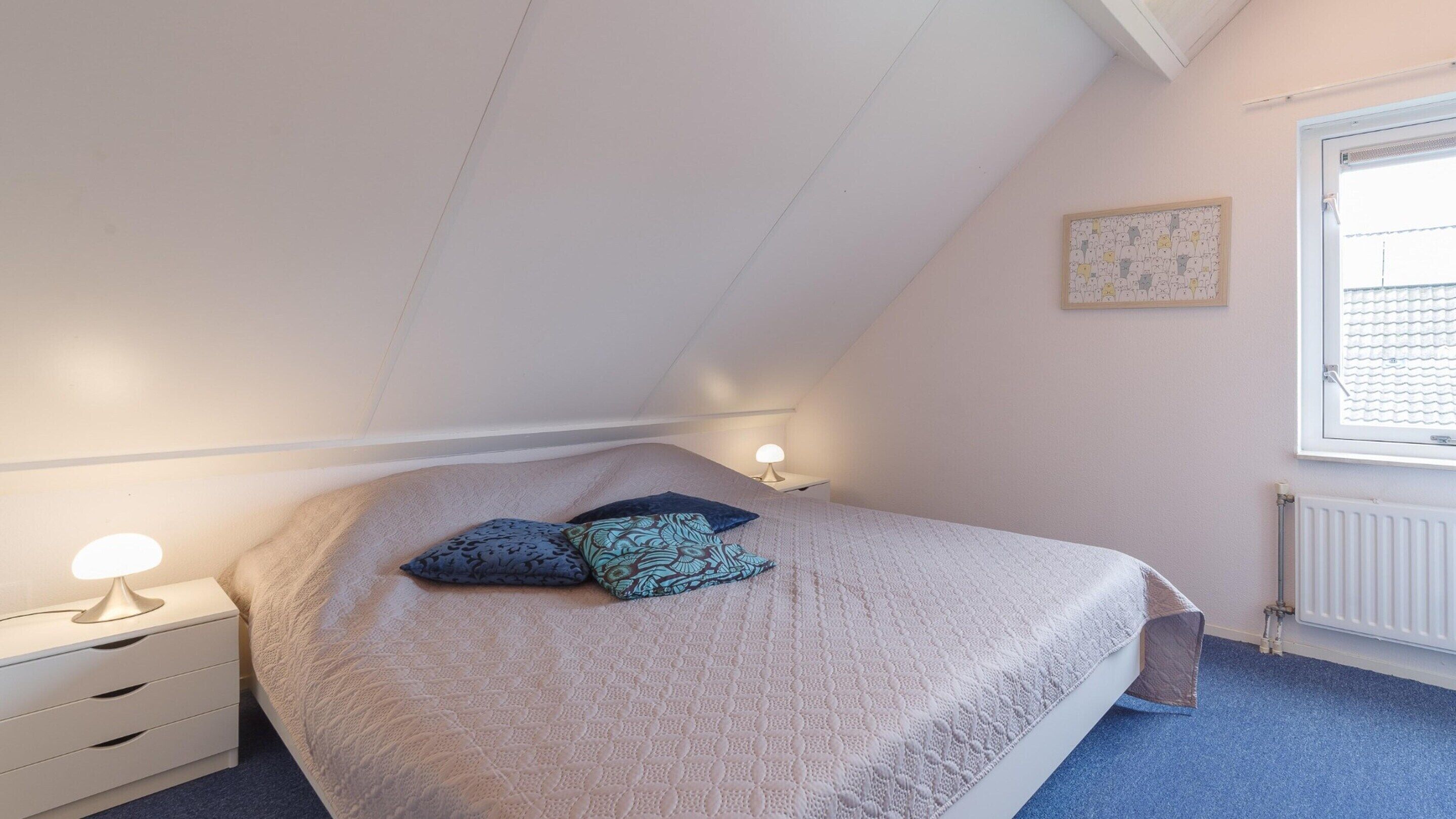 A bedroom with a sloped ceiling, a bed with a patterned bedspread and two pillows, bedside lamps, a window with blinds, and a radiator.
