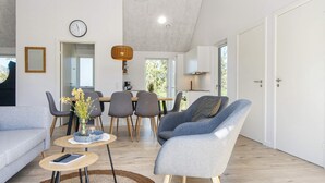 Un salon moderne comprenant un canapé, un fauteuil, une table à manger et des chaises. On y trouve également un coin cuisine avec des placards et une horloge au mur.