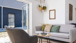 Un salon moderne avec un canapé gris, une table basse ronde et un fauteuil gris. On aperçoit une porte vitrée donnant sur l’extérieur, et une plante suspendue.