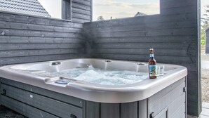 Un bain à remous avec une bouteille de bière et des verres, entouré de murs en bois et d’une fenêtre.