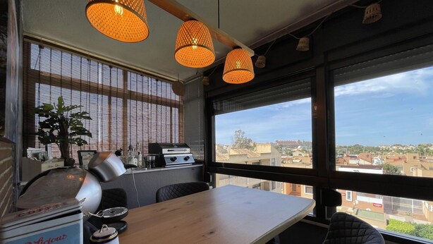 A cozy dining area with a wooden table, black cushioned chairs, a coffee station with a coffee can, and a view of the city through large windows.