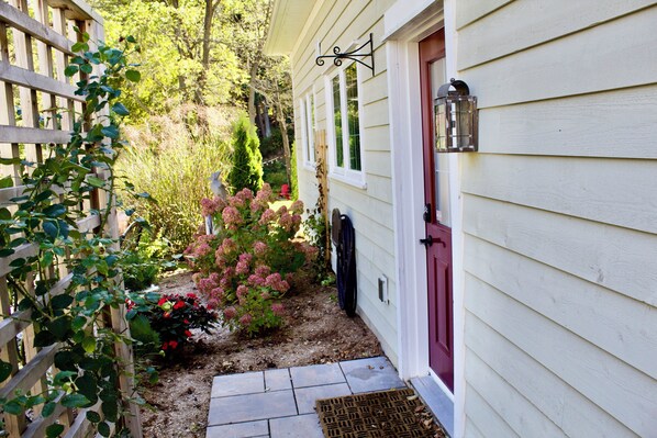 Une maison avec une porte rouge, une clôture en bois et un jardin où poussent des fleurs roses.