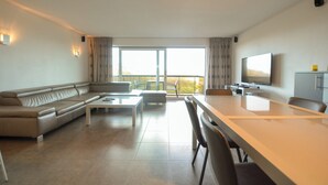 Una sala de estar moderna con sofá, mesa de centro y mesa de comedor. Hay un televisor montado en la pared y un balcón con cortinas.