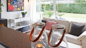 A modern living room with a glass-top coffee table, a white sofa, and a large window offering a view of a garden. - Tranquil Retreat on Eno - By Traum Ferienwohnungen (Karrebæksminde)
