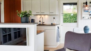 A modern kitchen with white cabinets, a built-in island with a sink, and a grey upholstered chair with a white cushion. - Tranquil Retreat on Eno - By Traum Ferienwohnungen (Karrebæksminde)