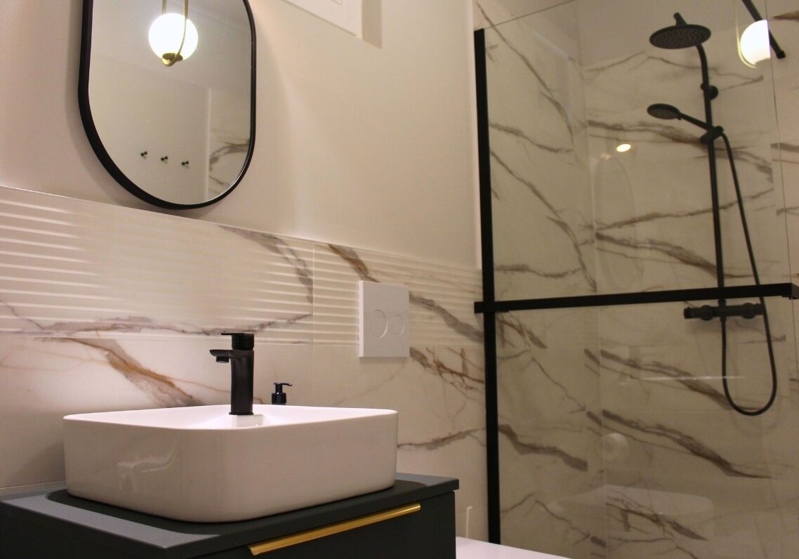 Une salle de bain avec un lavabo rond blanc, un robinet noir, un miroir à cadre noir et une douche à parois de verre, dotée d’un flexible noir.