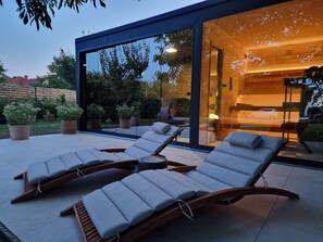 A modern outdoor lounge area with two sun loungers and a small table, surrounded by potted plants and a glass wall offering a view of the interior.